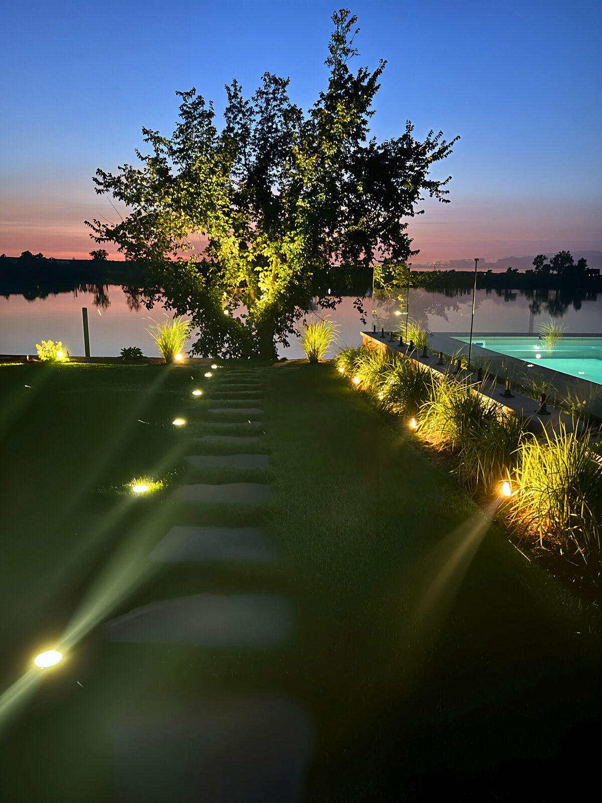 Path and trees uplit beside water at dusk